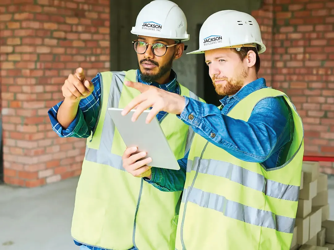 Two construction workers in safety gear discuss plans via a tablet in a brick-walled building under construction